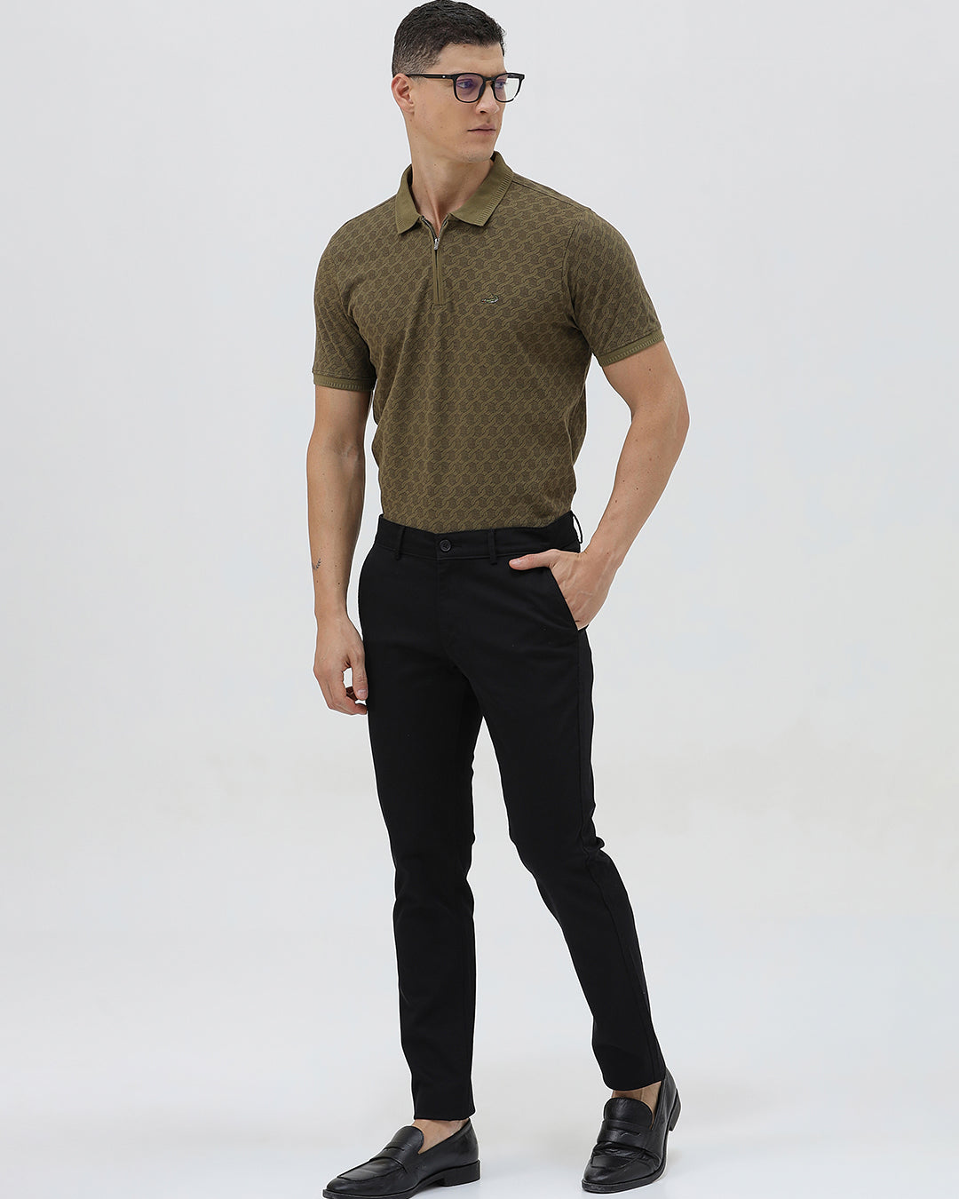 A man wearing a short-sleeve polo shirt with a subtle geometric monogram pattern in olive green and brown, paired with black trousers, against a light gray studio background.