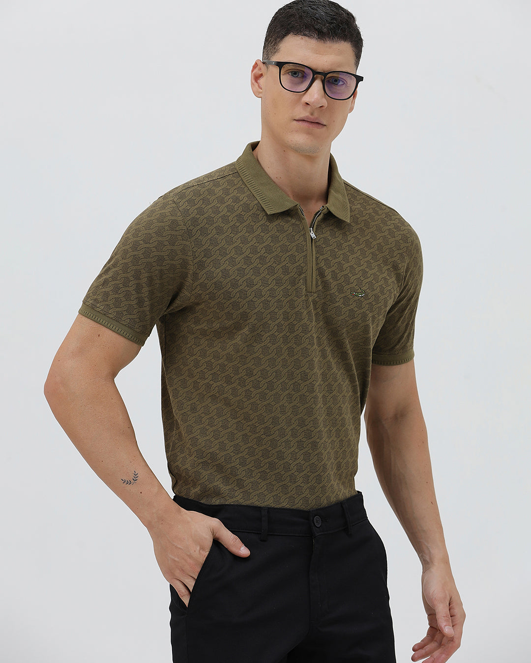 A man wearing a short-sleeve polo shirt with a subtle geometric monogram pattern in olive green and brown, paired with black trousers, against a light gray studio background.