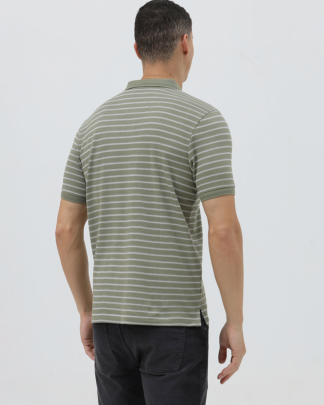 A man wearing a short-sleeve polo shirt with thin horizontal olive green and white stripes, paired with dark gray pants, standing against a light gray studio background.