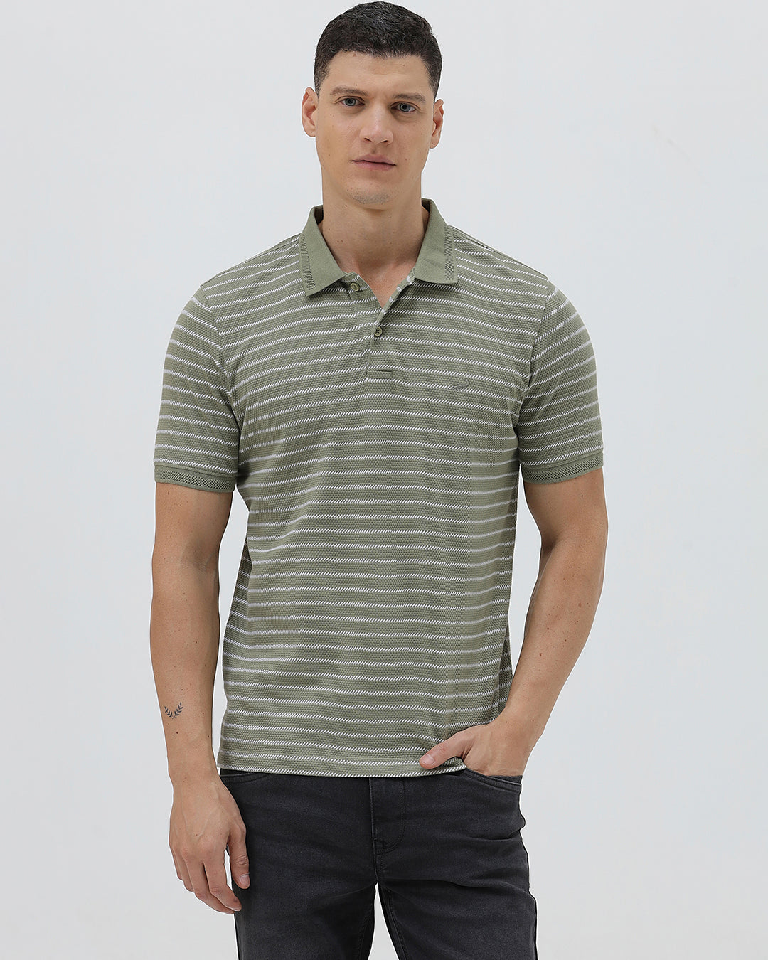 A man wearing a short-sleeve polo shirt with thin horizontal olive green and white stripes, paired with dark gray pants, standing against a light gray studio background.