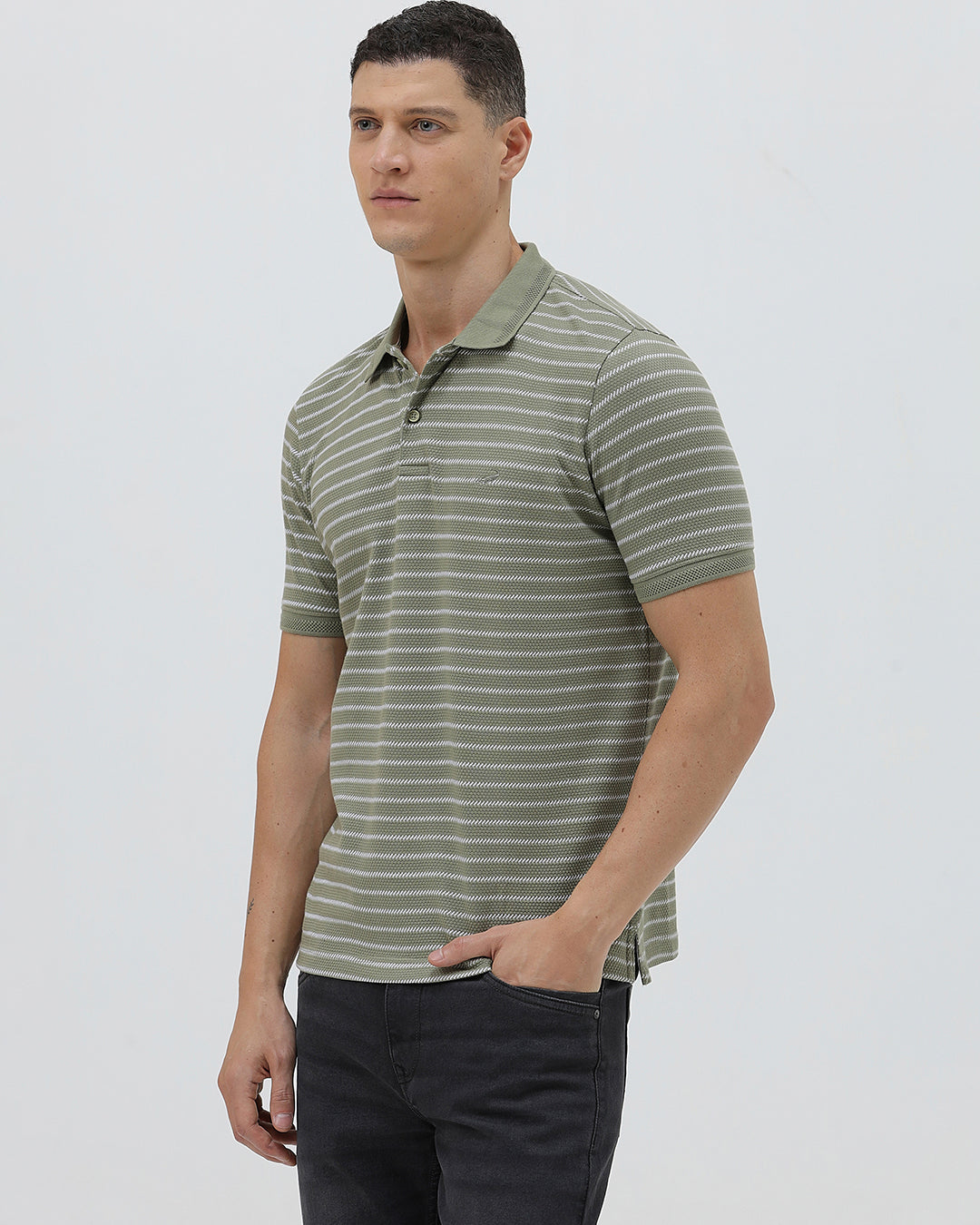 A man wearing a short-sleeve polo shirt with thin horizontal olive green and white stripes, paired with dark gray pants, standing against a light gray studio background.