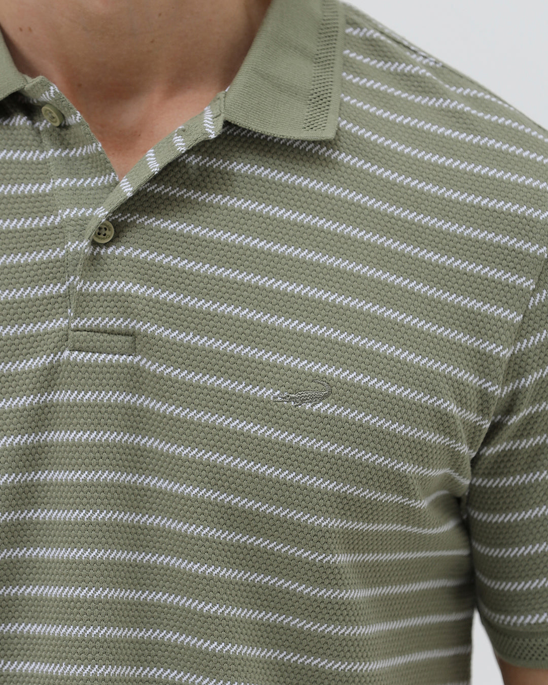 A man wearing a short-sleeve polo shirt with thin horizontal olive green and white stripes, paired with dark gray pants, standing against a light gray studio background.