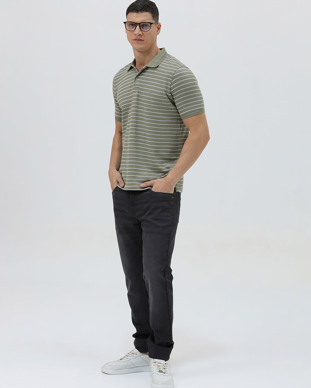 A man wearing a short-sleeve polo shirt with thin horizontal olive green and white stripes, paired with dark gray pants, standing against a light gray studio background.