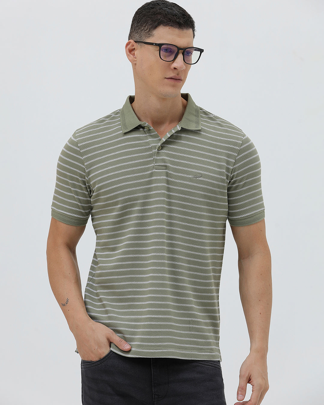 A man wearing a short-sleeve polo shirt with thin horizontal olive green and white stripes, paired with dark gray pants, standing against a light gray studio background.