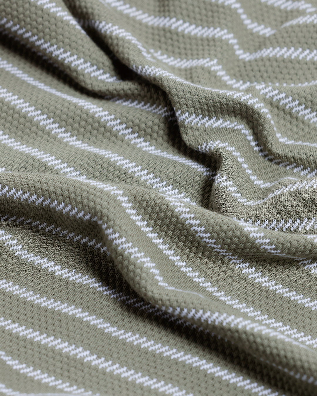A man wearing a short-sleeve polo shirt with thin horizontal olive green and white stripes, paired with dark gray pants, standing against a light gray studio background.