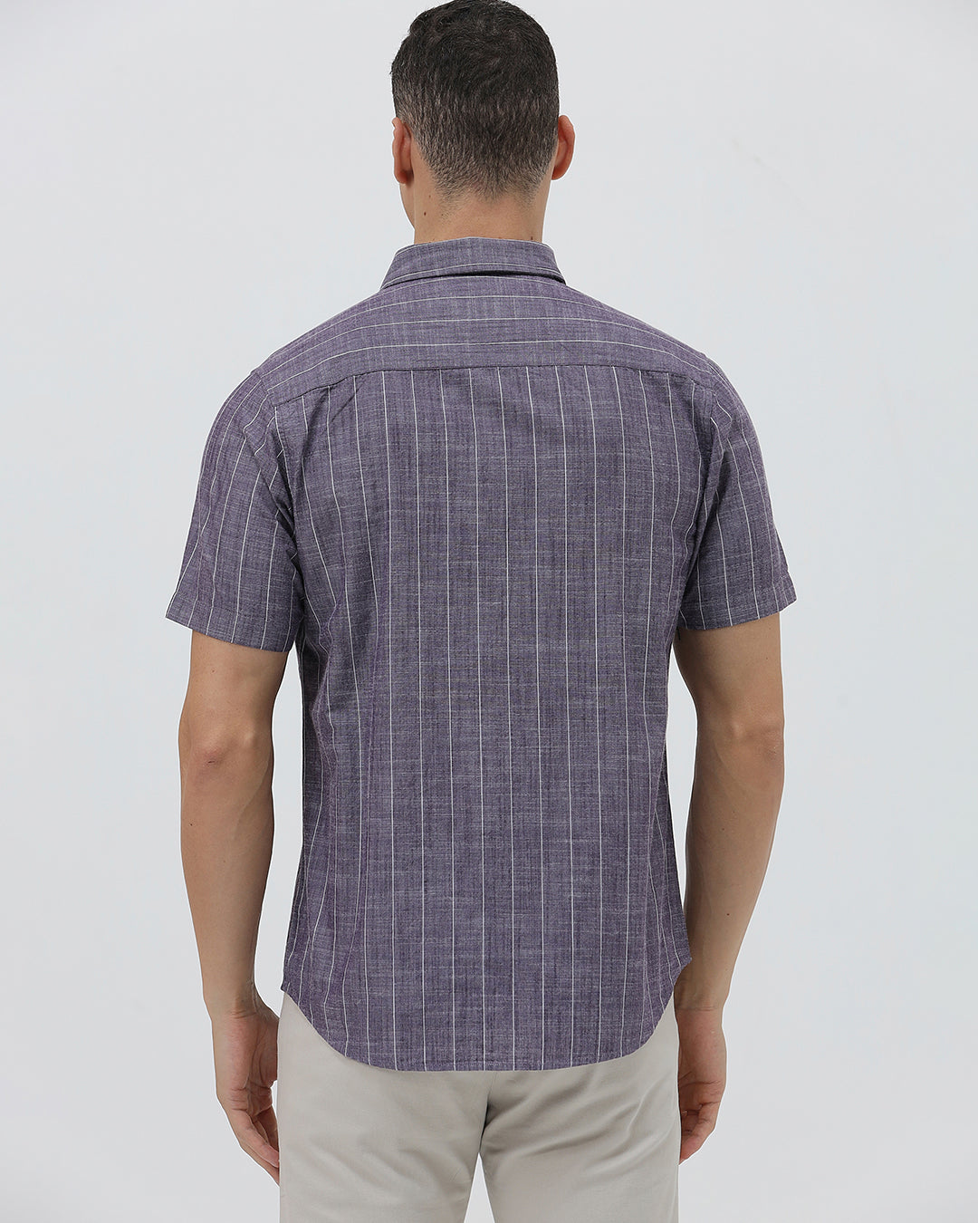 A man wearing a short-sleeve button-up shirt with a purple and white vertical stripe pattern, paired with light beige trousers, standing against a light gray studio background.