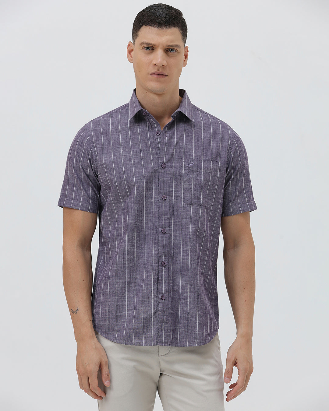 A man wearing a short-sleeve button-up shirt with a purple and white vertical stripe pattern, paired with light beige trousers, standing against a light gray studio background.