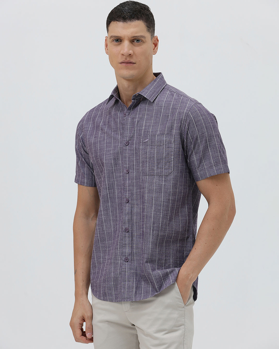 A man wearing a short-sleeve button-up shirt with a purple and white vertical stripe pattern, paired with light beige trousers, standing against a light gray studio background.