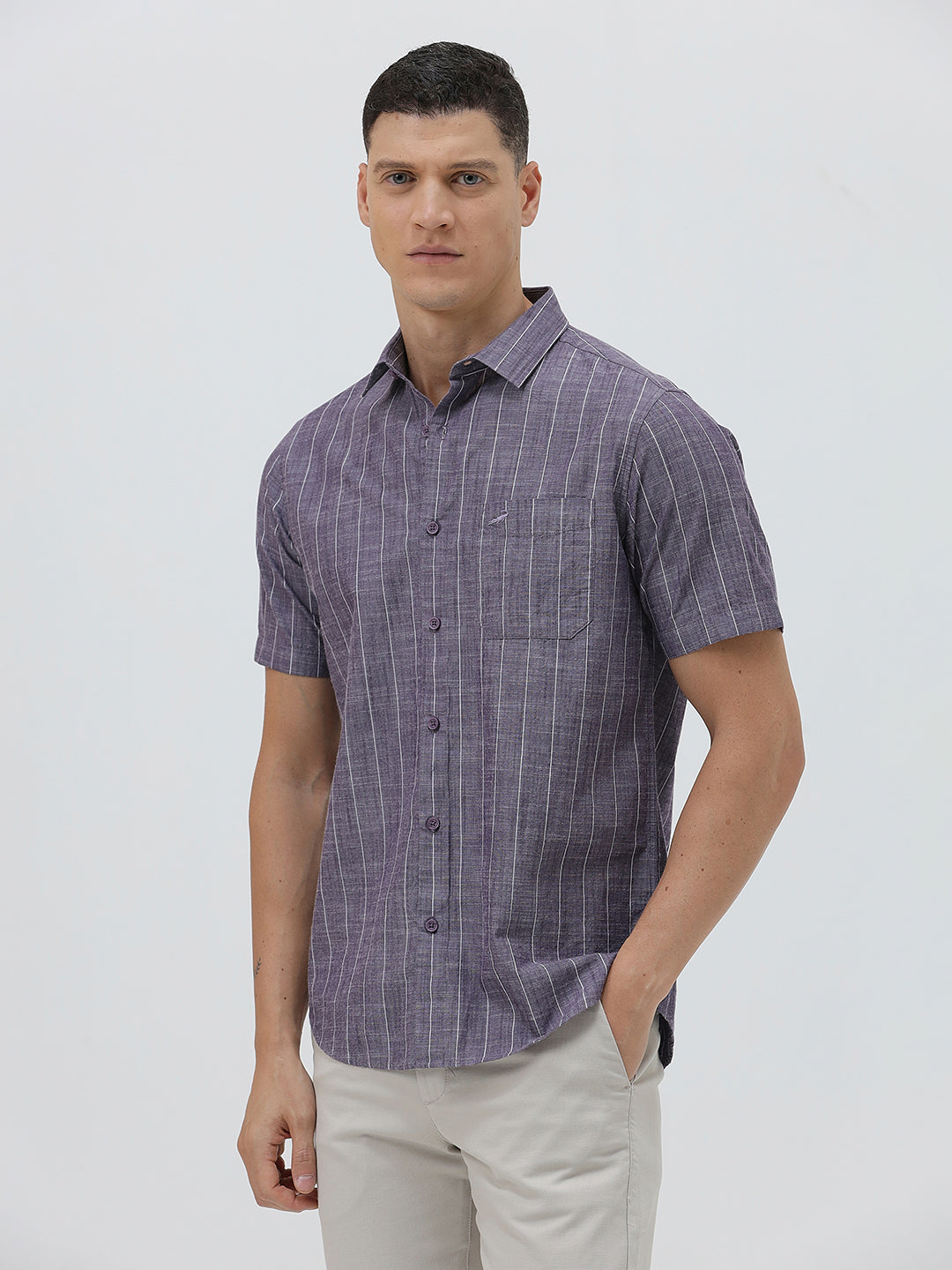 A man wearing a short-sleeve button-up shirt with a purple and white vertical stripe pattern, paired with light beige trousers, standing against a light gray studio background.