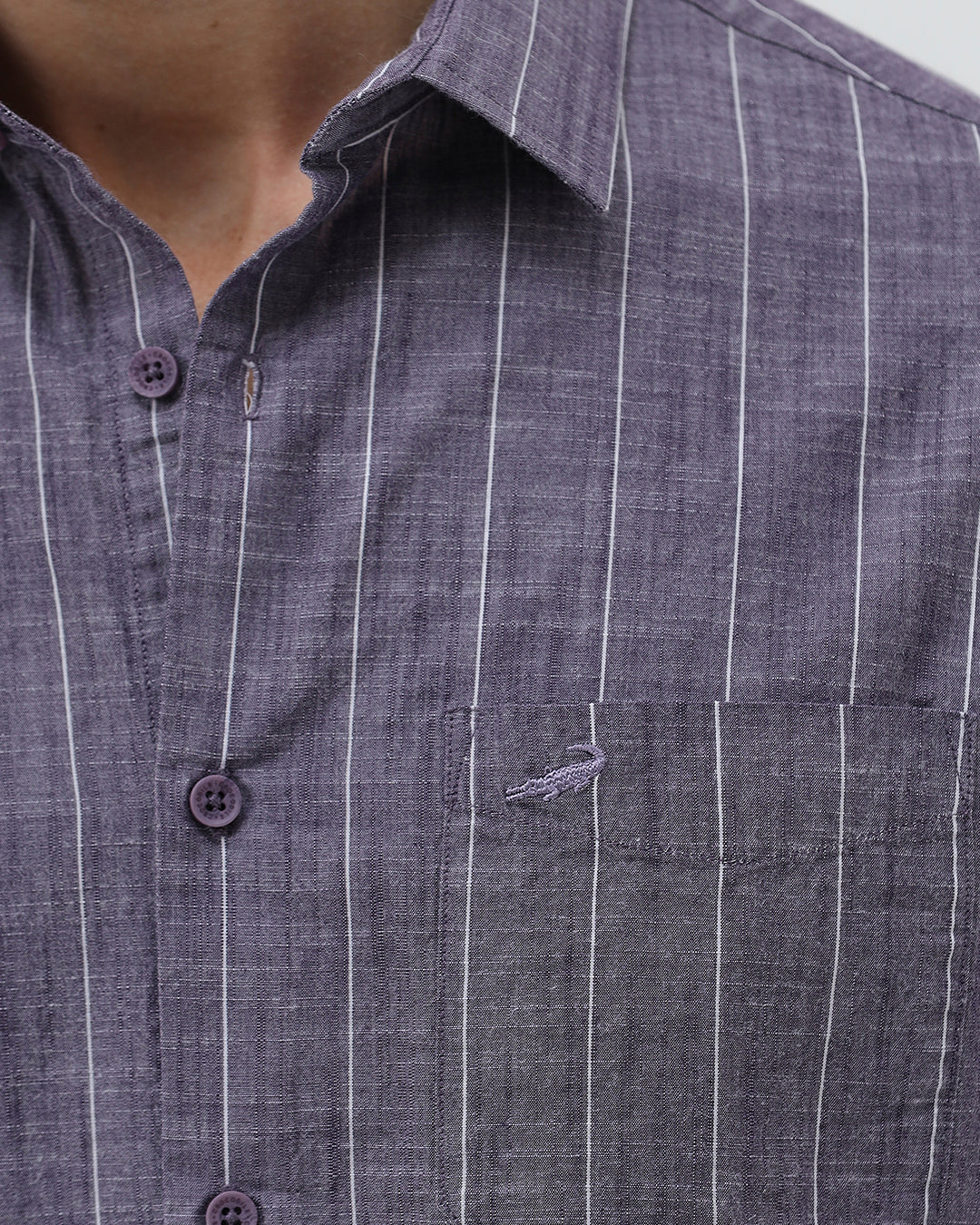 A man wearing a short-sleeve button-up shirt with a purple and white vertical stripe pattern, paired with light beige trousers, standing against a light gray studio background.