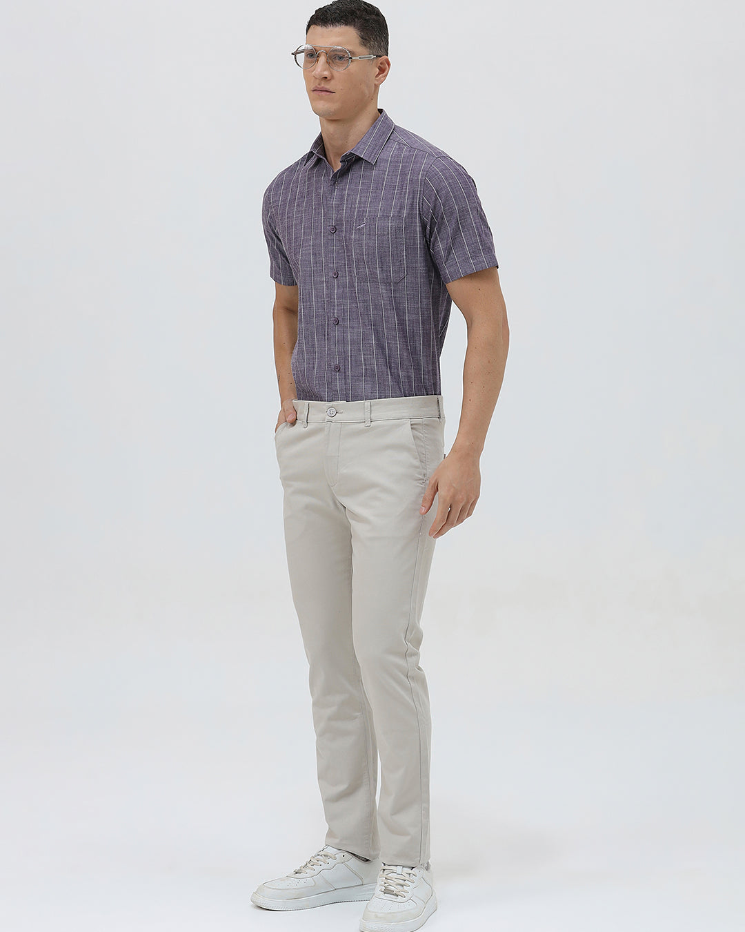 A man wearing a short-sleeve button-up shirt with a purple and white vertical stripe pattern, paired with light beige trousers, standing against a light gray studio background.
