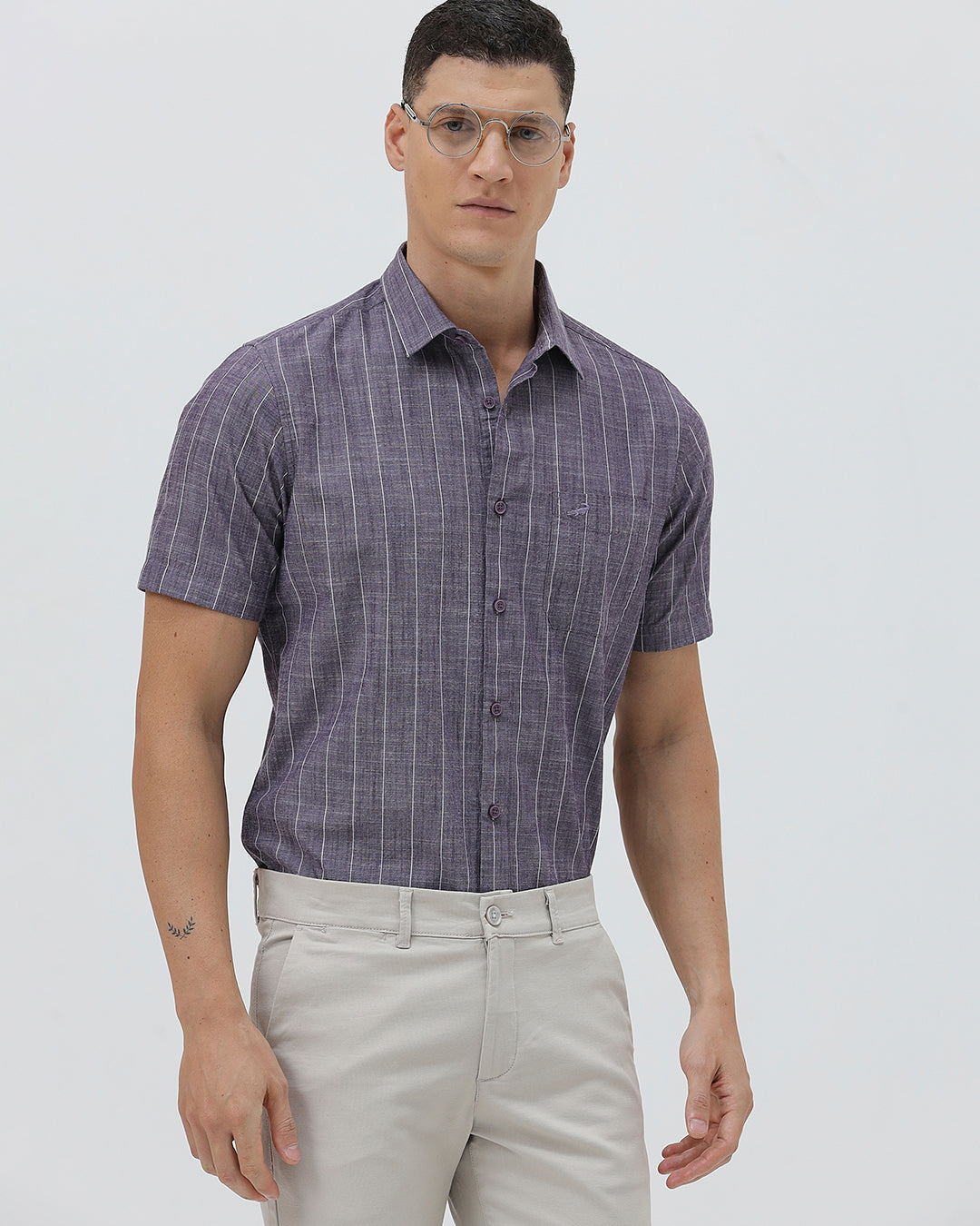 A man wearing a short-sleeve button-up shirt with a purple and white vertical stripe pattern, paired with light beige trousers, standing against a light gray studio background.