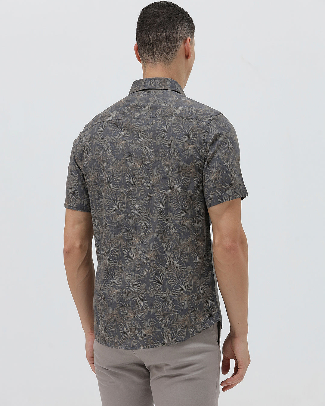 A man wearing a short-sleeve button-up shirt with a dark gray and brown palm leaf pattern, paired with light gray trousers, standing against a light gray background.