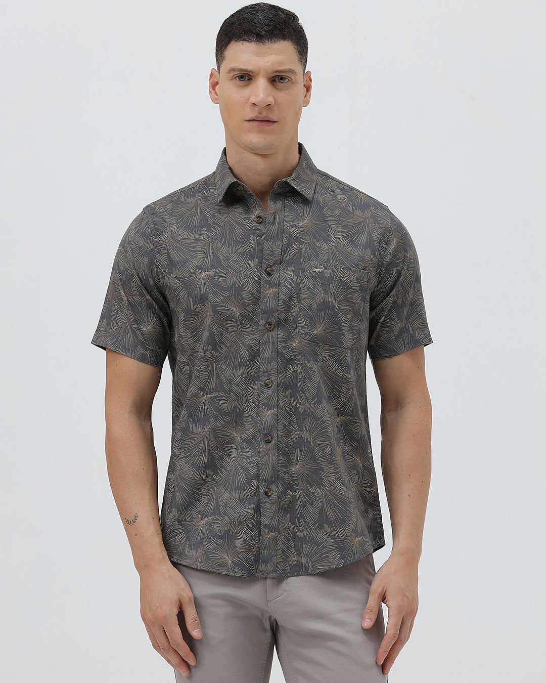 A man wearing a short-sleeve button-up shirt with a dark gray and brown palm leaf pattern, paired with light gray trousers, standing against a light gray background.