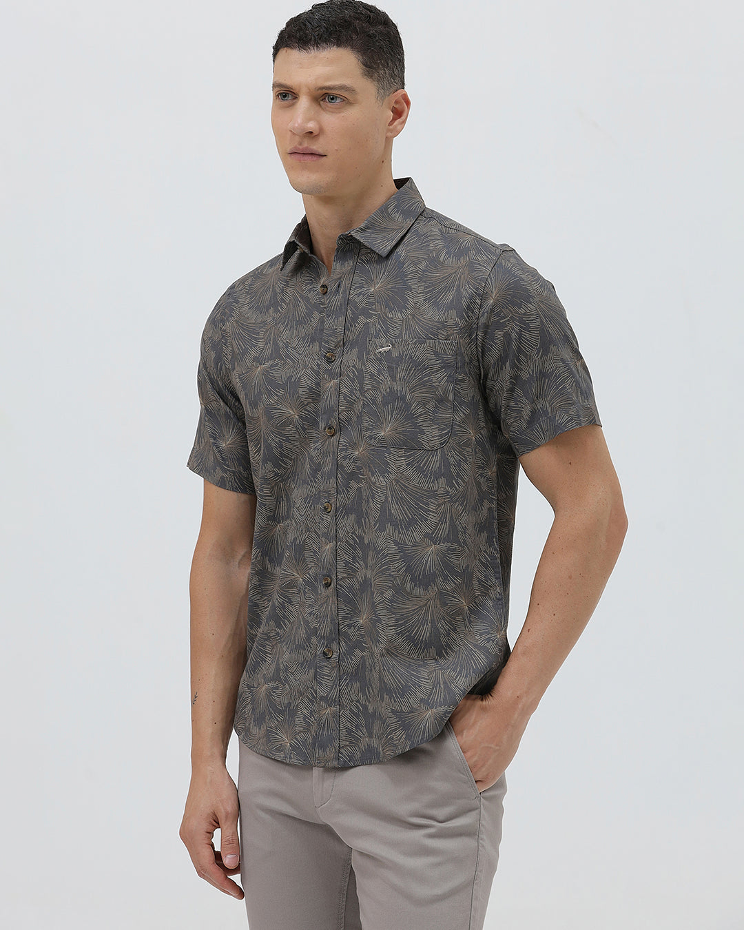 A man wearing a short-sleeve button-up shirt with a dark gray and brown palm leaf pattern, paired with light gray trousers, standing against a light gray background.