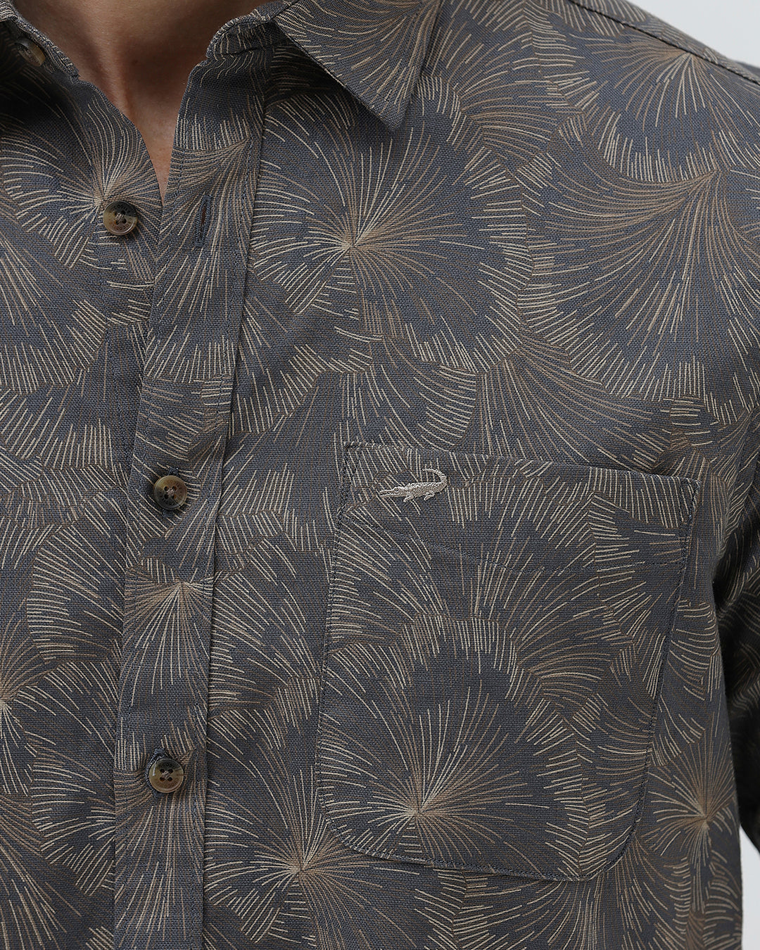A man wearing a short-sleeve button-up shirt with a dark gray and brown palm leaf pattern, paired with light gray trousers, standing against a light gray background.