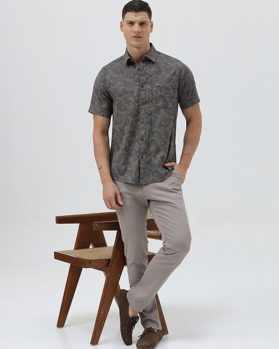 A man wearing a short-sleeve button-up shirt with a dark gray and brown palm leaf pattern, paired with light gray trousers, standing against a light gray background.
