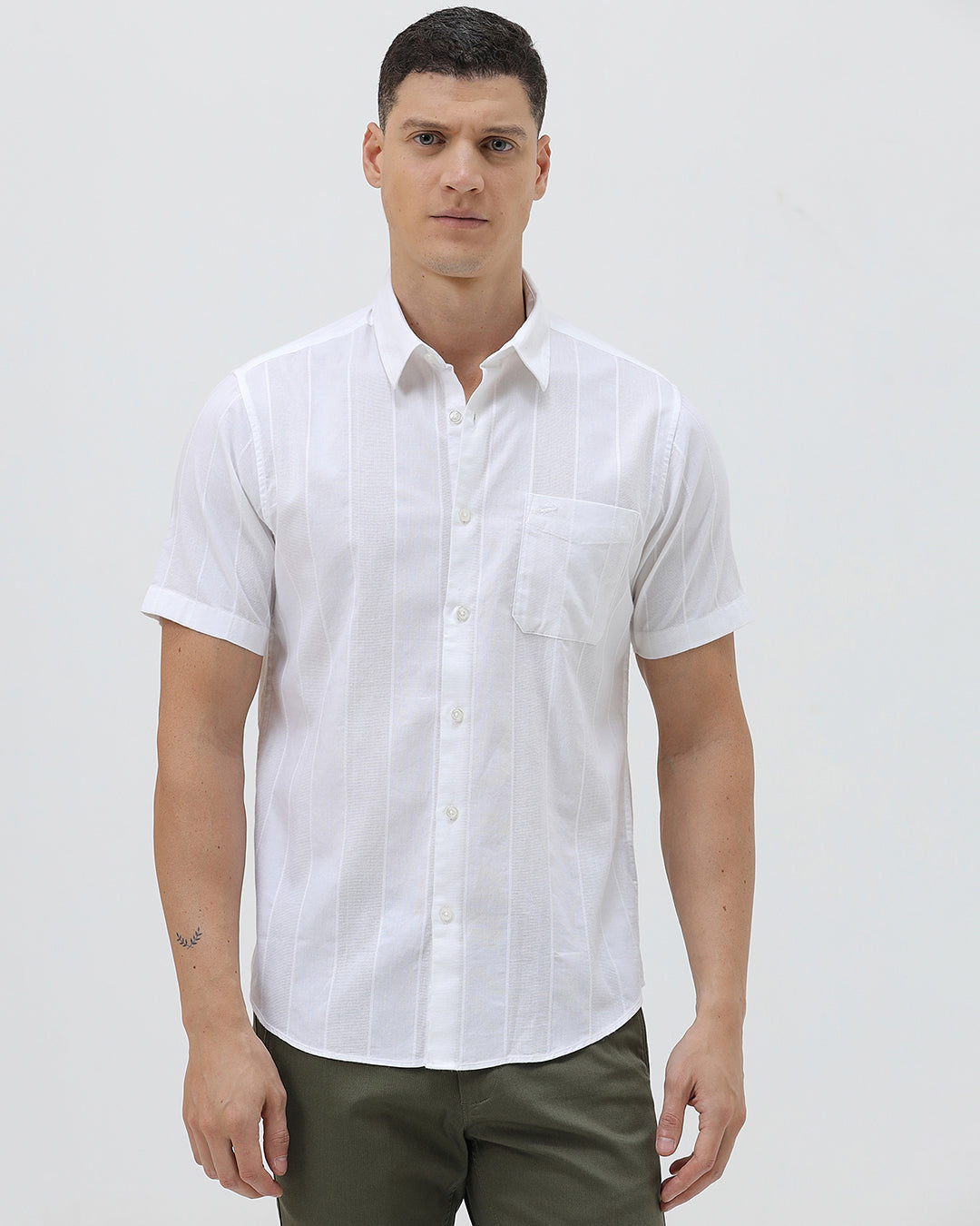 A man wearing a white short-sleeve button-up shirt with subtle vertical stripes and olive green trousers, standing against a plain light gray background.