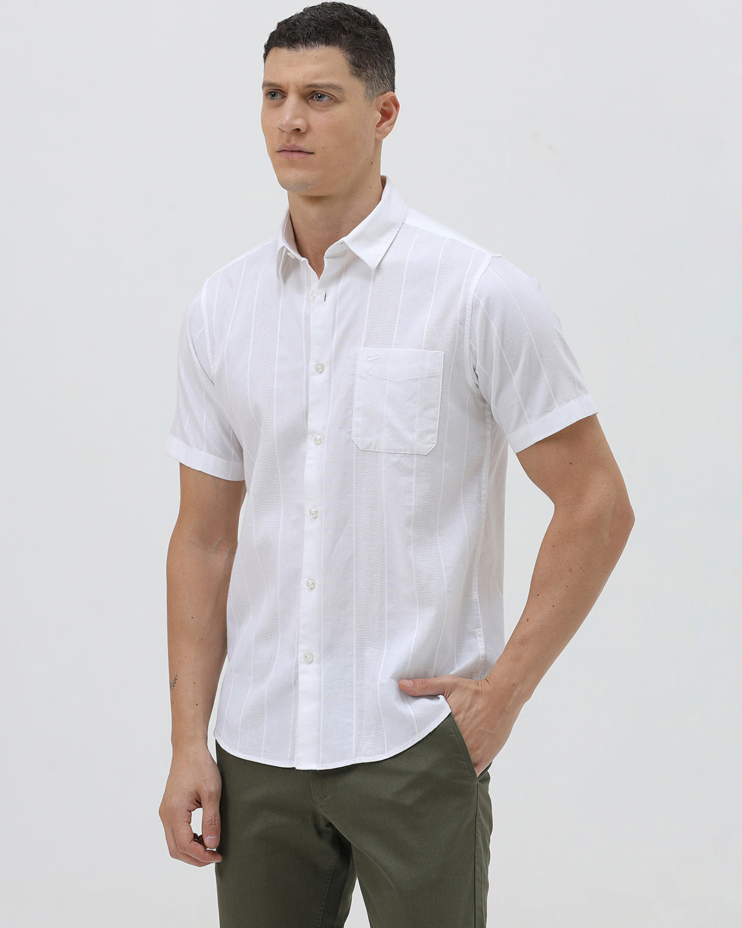 A man wearing a white short-sleeve button-up shirt with subtle vertical stripes and olive green trousers, standing against a plain light gray background.