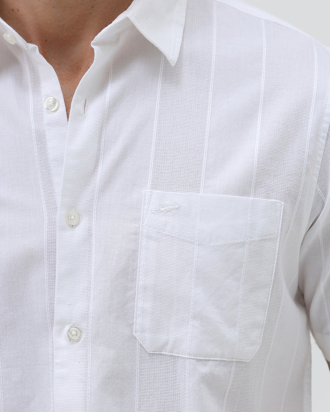 A man wearing a white short-sleeve button-up shirt with subtle vertical stripes and olive green trousers, standing against a plain light gray background.