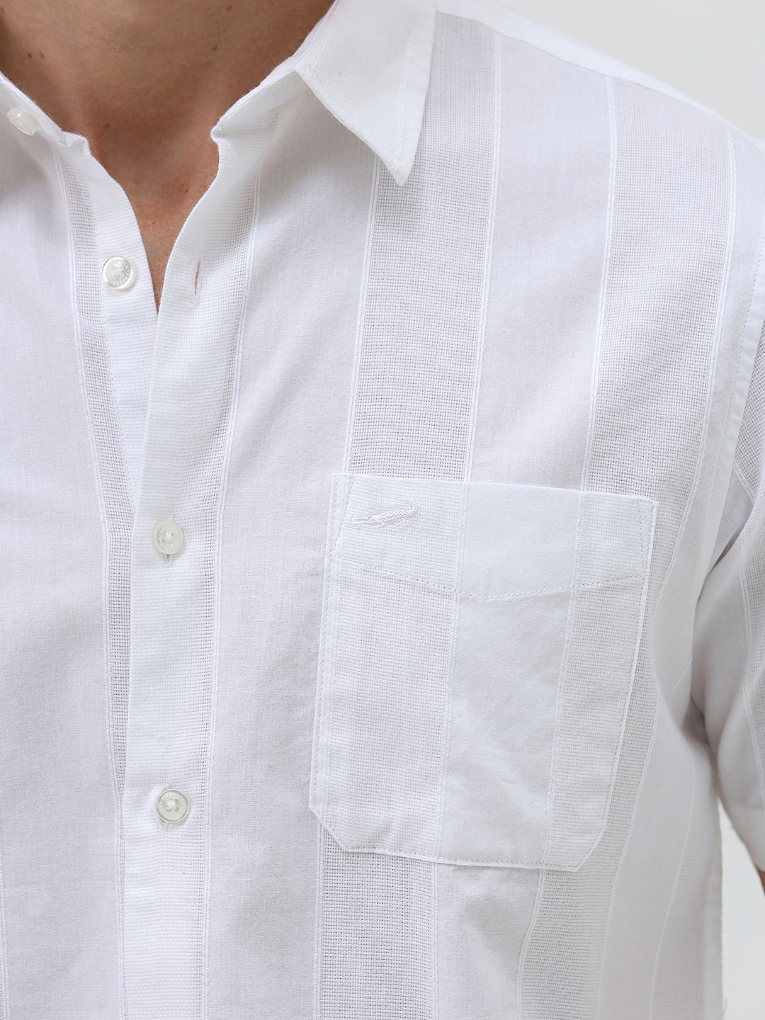 A man wearing a white short-sleeve button-up shirt with subtle vertical stripes and olive green trousers, standing against a plain light gray background.