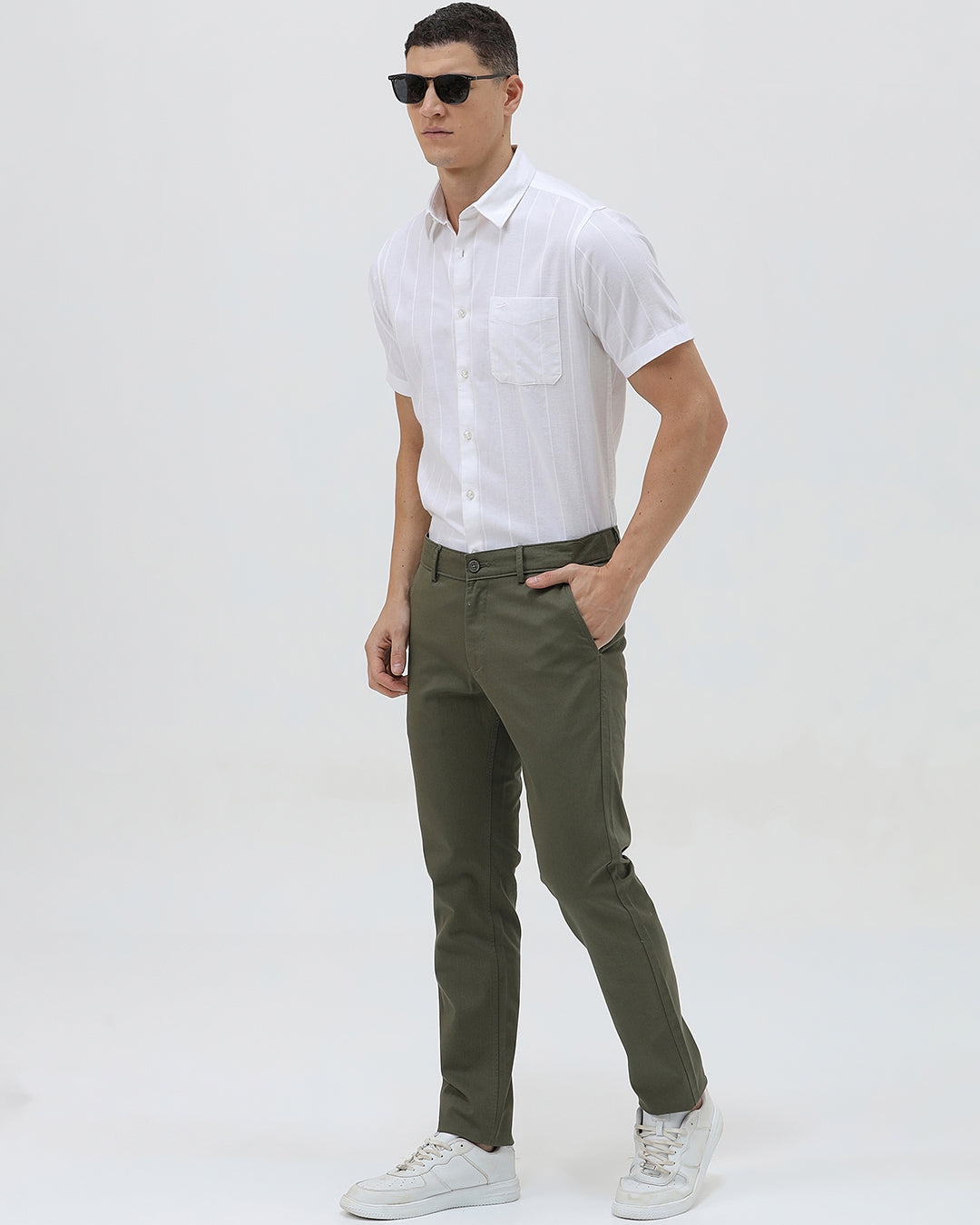 A man wearing a white short-sleeve button-up shirt with subtle vertical stripes and olive green trousers, standing against a plain light gray background.