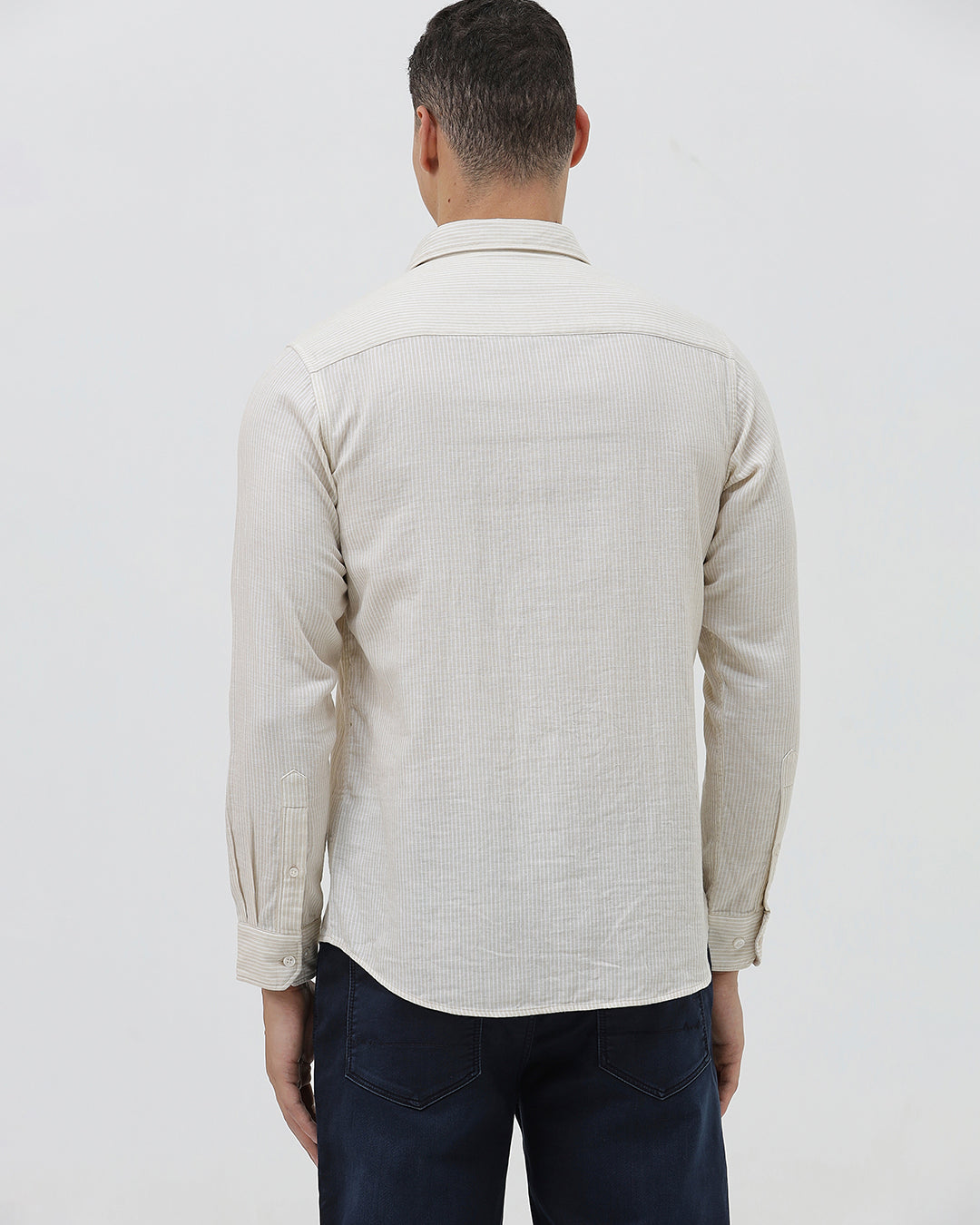 A man wearing a light beige linen long-sleeve button-up shirt with a chest pocket, paired with dark blue jeans, standing against a plain white studio background.