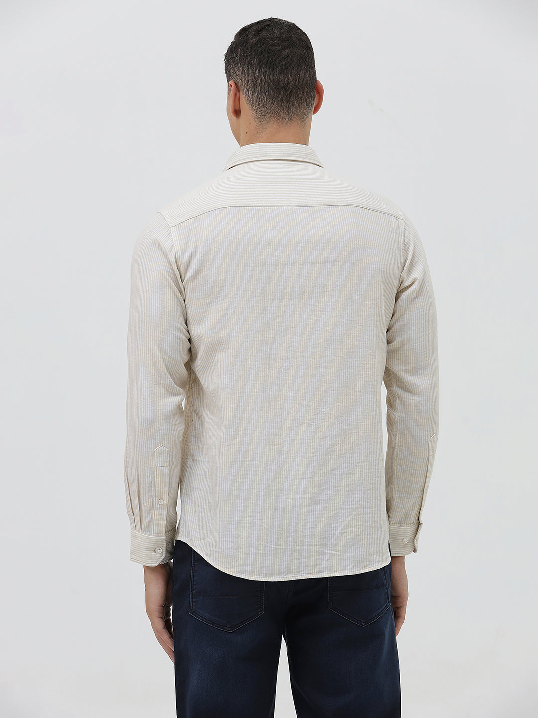 A man wearing a light beige linen long-sleeve button-up shirt with a chest pocket, paired with dark blue jeans, standing against a plain white studio background.