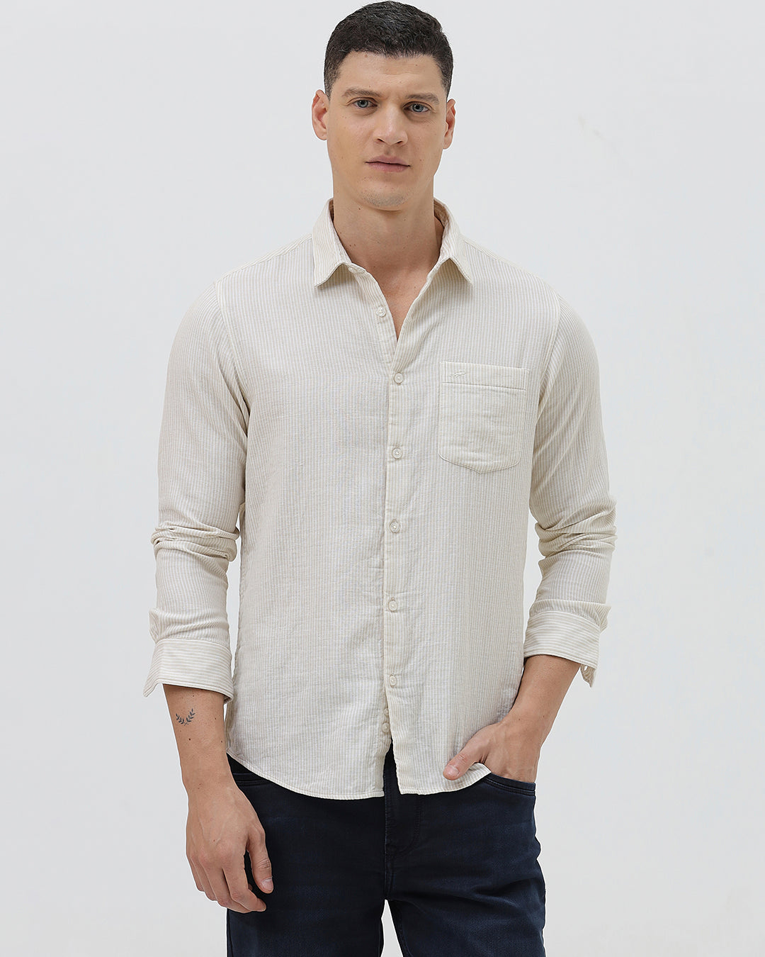 A man wearing a light beige linen long-sleeve button-up shirt with a chest pocket, paired with dark blue jeans, standing against a plain white studio background.