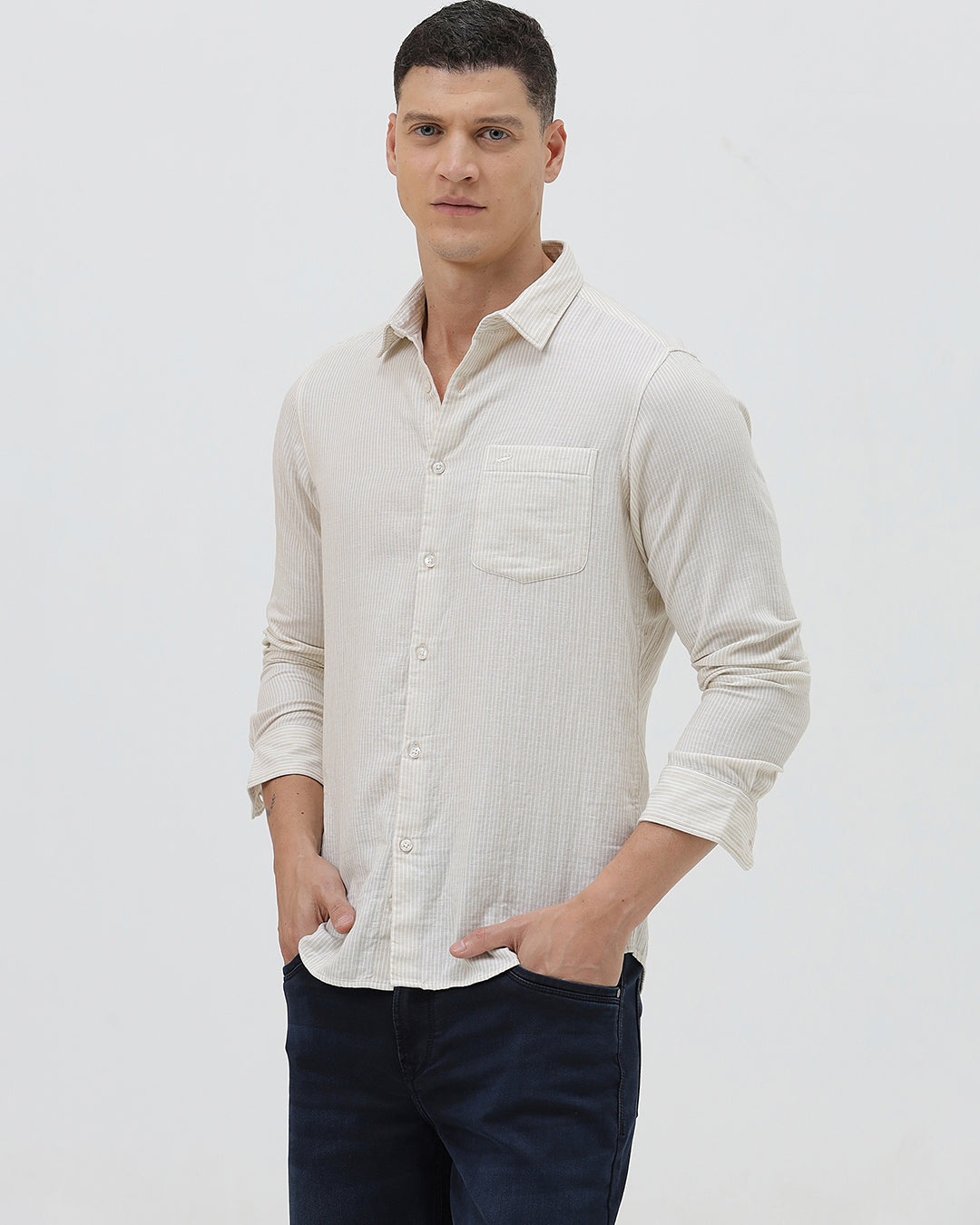 A man wearing a light beige linen long-sleeve button-up shirt with a chest pocket, paired with dark blue jeans, standing against a plain white studio background.
