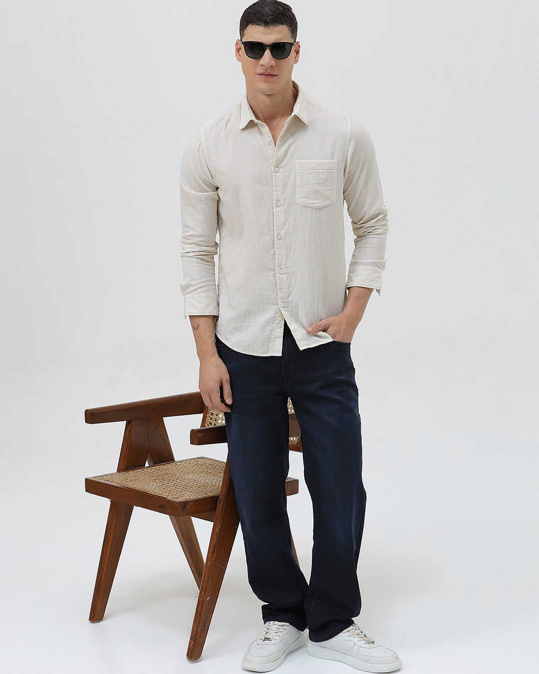 A man wearing a light beige linen long-sleeve button-up shirt with a chest pocket, paired with dark blue jeans, standing against a plain white studio background.