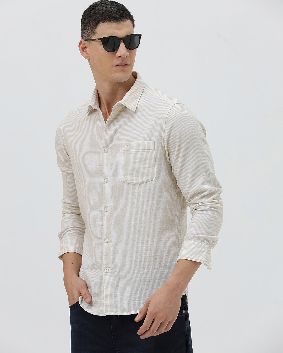 A man wearing a light beige linen long-sleeve button-up shirt with a chest pocket, paired with dark blue jeans, standing against a plain white studio background.