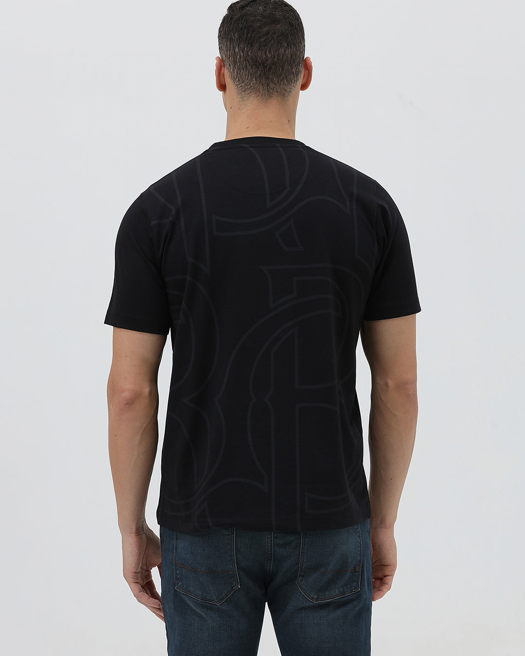 A man wearing a black short-sleeve t-shirt with a subtle tonal geometric pattern and a small 'Gildan' logo on the left chest, paired with dark blue jeans, against a light gray studio background.