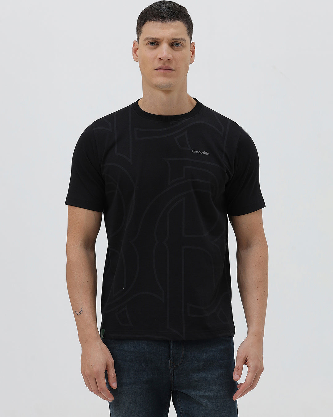 A man wearing a black short-sleeve t-shirt with a subtle tonal geometric pattern and a small 'Gildan' logo on the left chest, paired with dark blue jeans, against a light gray studio background.