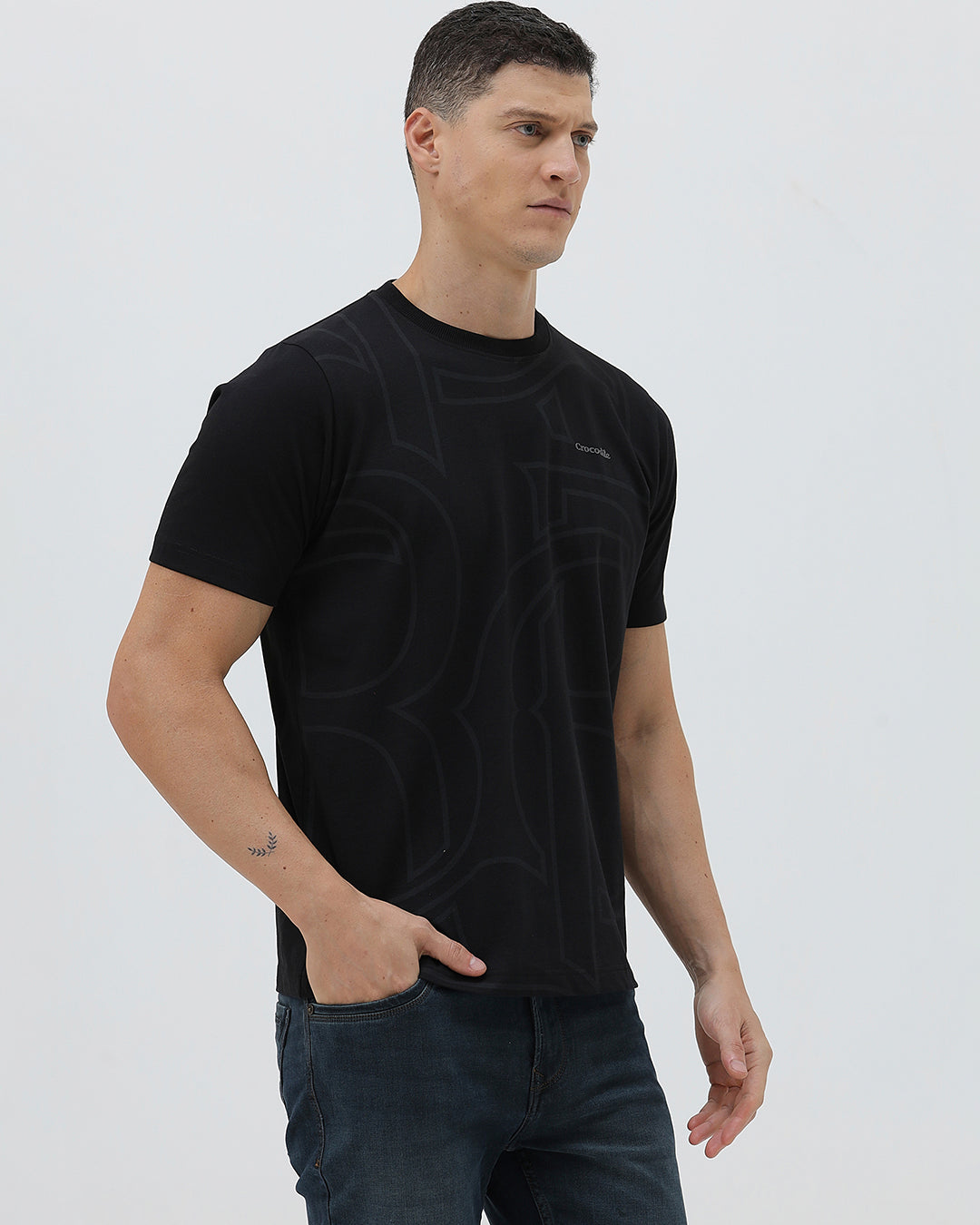 A man wearing a black short-sleeve t-shirt with a subtle tonal geometric pattern and a small 'Gildan' logo on the left chest, paired with dark blue jeans, against a light gray studio background.