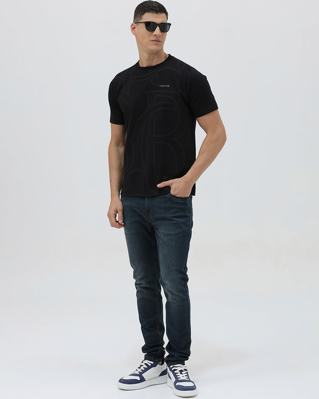 A man wearing a black short-sleeve t-shirt with a subtle tonal geometric pattern and a small 'Gildan' logo on the left chest, paired with dark blue jeans, against a light gray studio background.
