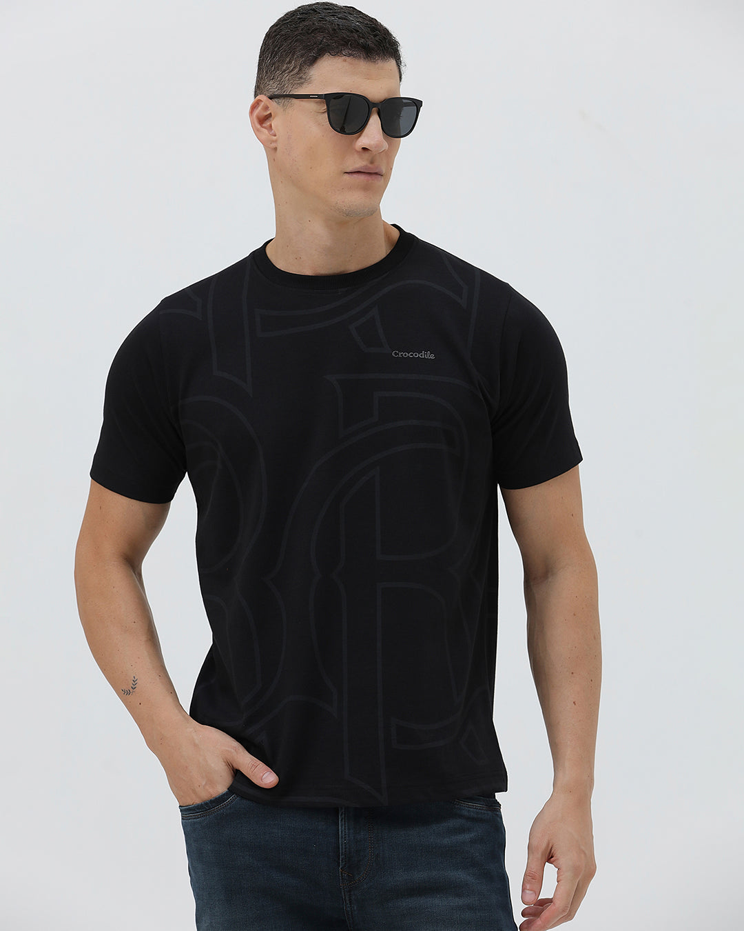 A man wearing a black short-sleeve t-shirt with a subtle tonal geometric pattern and a small 'Gildan' logo on the left chest, paired with dark blue jeans, against a light gray studio background.