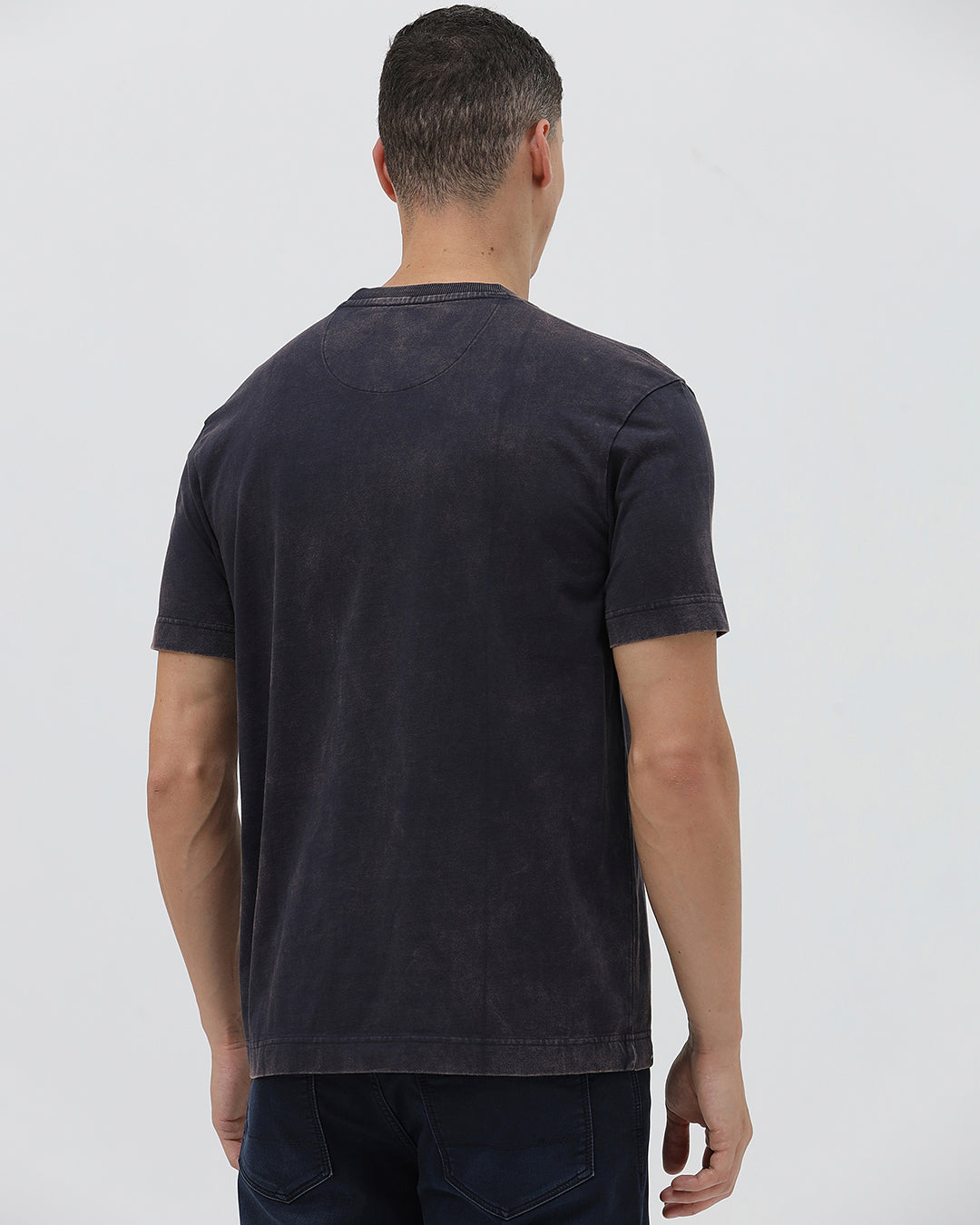 A man wearing a dark charcoal gray washed t-shirt with a blue graphic text print reading 'CROCODILE' on the chest, paired with dark blue jeans, against a light gray studio background.