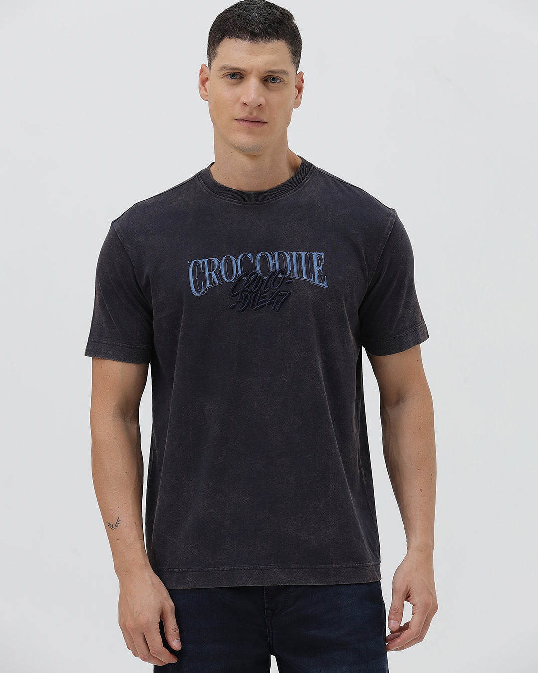 A man wearing a dark charcoal gray washed t-shirt with a blue graphic text print reading 'CROCODILE' on the chest, paired with dark blue jeans, against a light gray studio background.