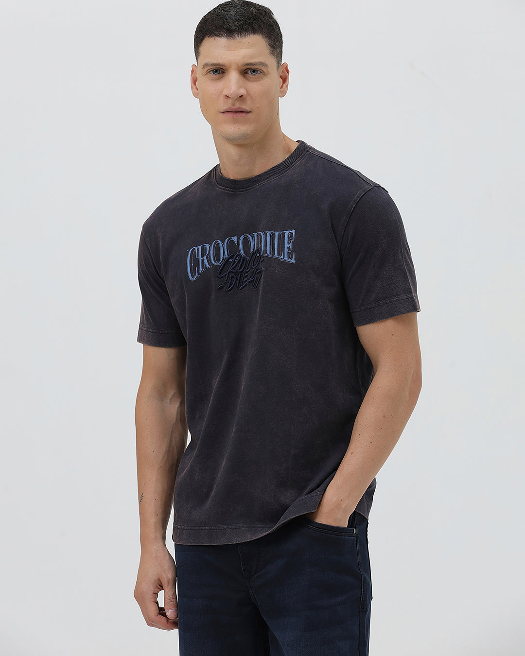 A man wearing a dark charcoal gray washed t-shirt with a blue graphic text print reading 'CROCODILE' on the chest, paired with dark blue jeans, against a light gray studio background.
