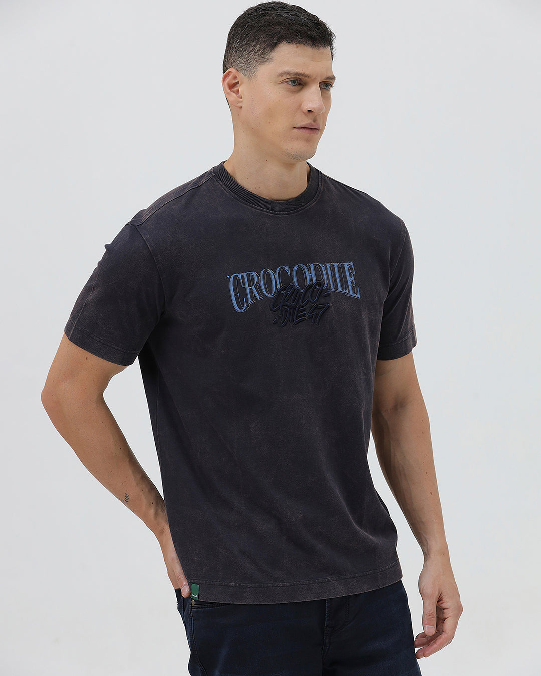 A man wearing a dark charcoal gray washed t-shirt with a blue graphic text print reading 'CROCODILE' on the chest, paired with dark blue jeans, against a light gray studio background.