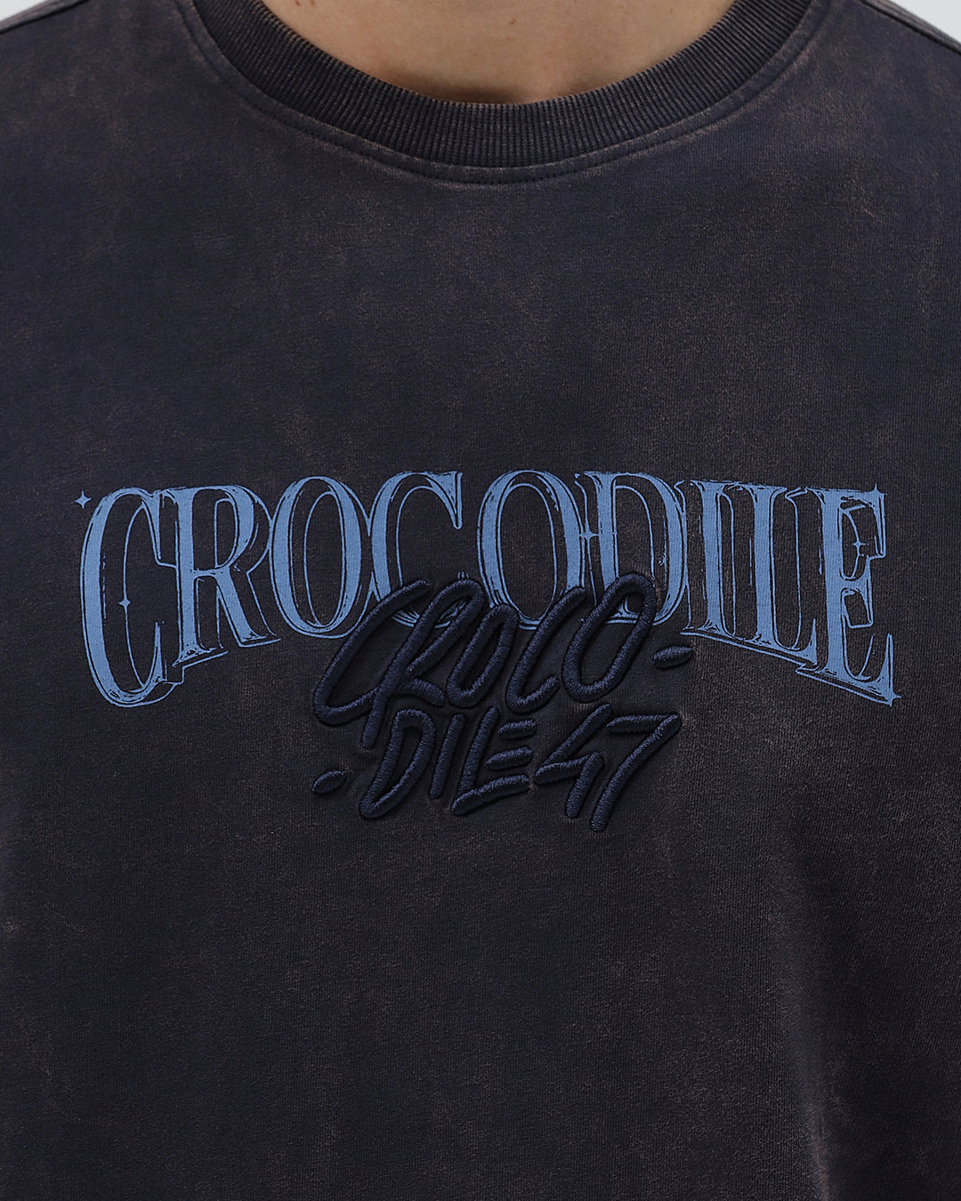 A man wearing a dark charcoal gray washed t-shirt with a blue graphic text print reading 'CROCODILE' on the chest, paired with dark blue jeans, against a light gray studio background.
