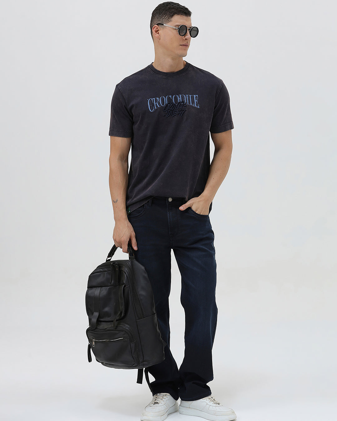 A man wearing a dark charcoal gray washed t-shirt with a blue graphic text print reading 'CROCODILE' on the chest, paired with dark blue jeans, against a light gray studio background.