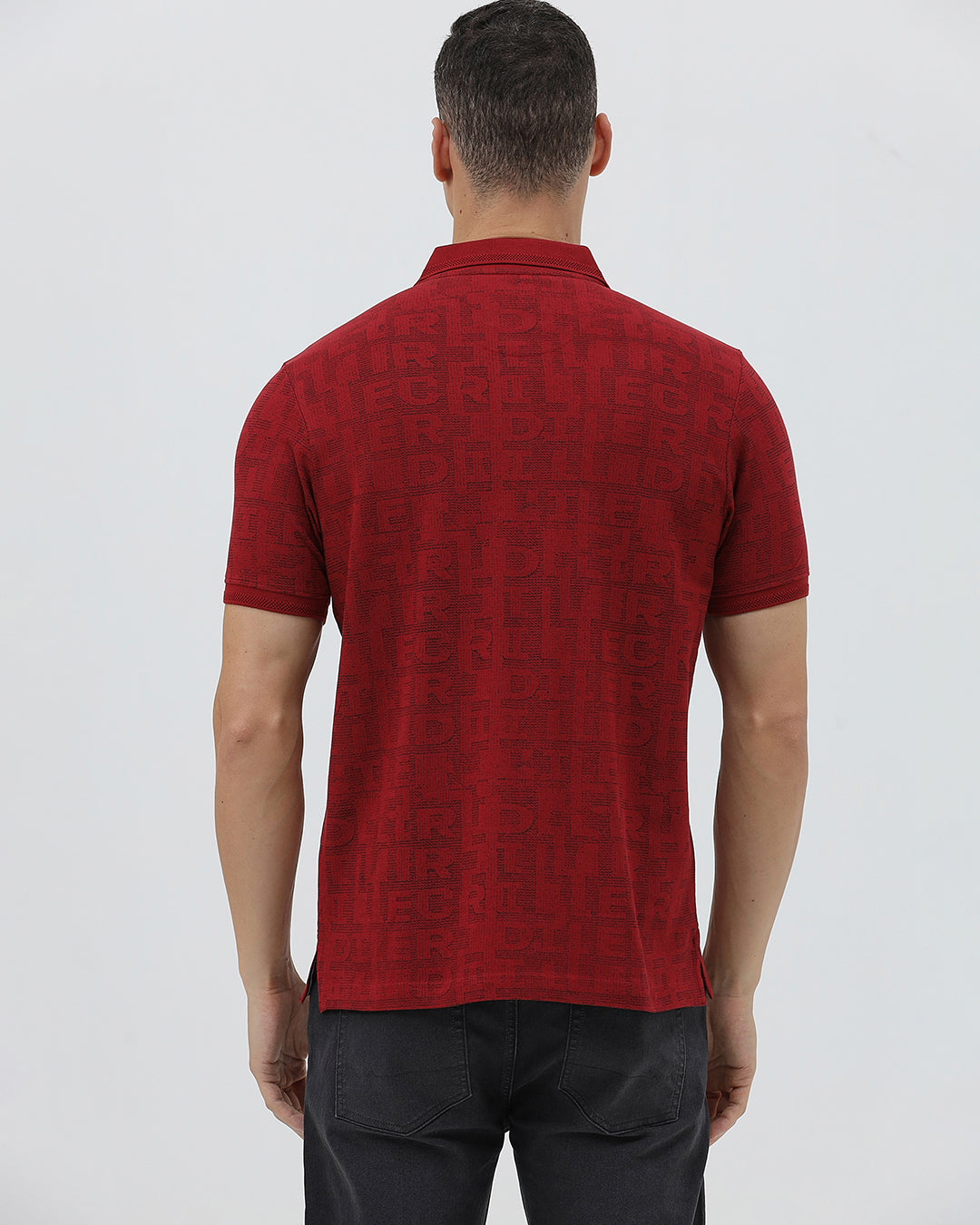 A man wearing a red short-sleeve polo shirt with a subtle tonal monogram pattern, paired with dark gray jeans, standing against a light gray studio background.