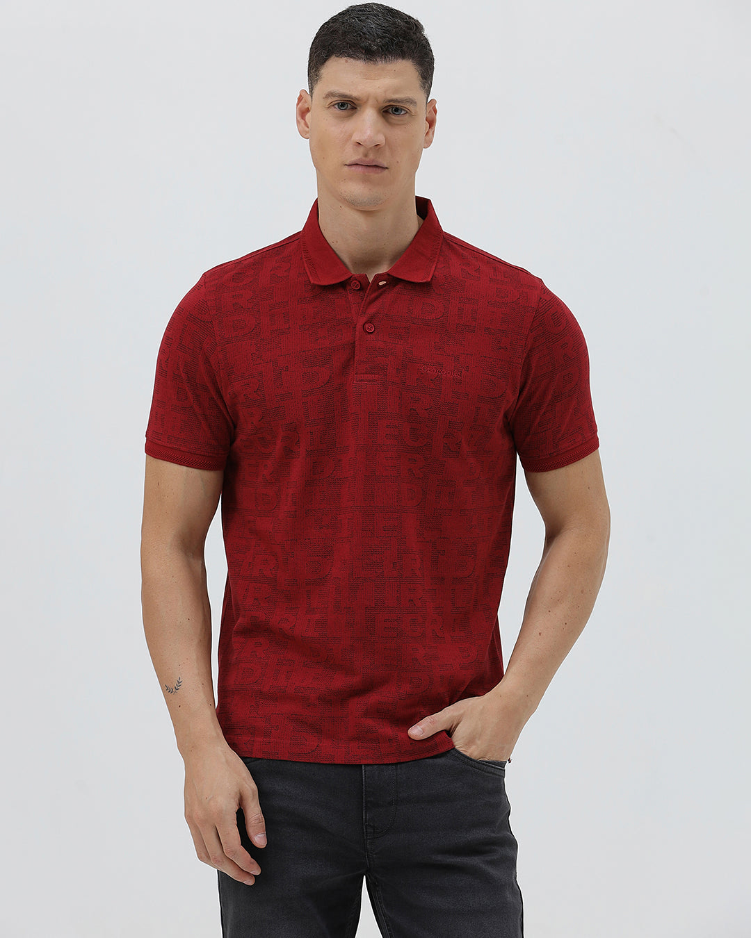 A man wearing a red short-sleeve polo shirt with a subtle tonal monogram pattern, paired with dark gray jeans, standing against a light gray studio background.