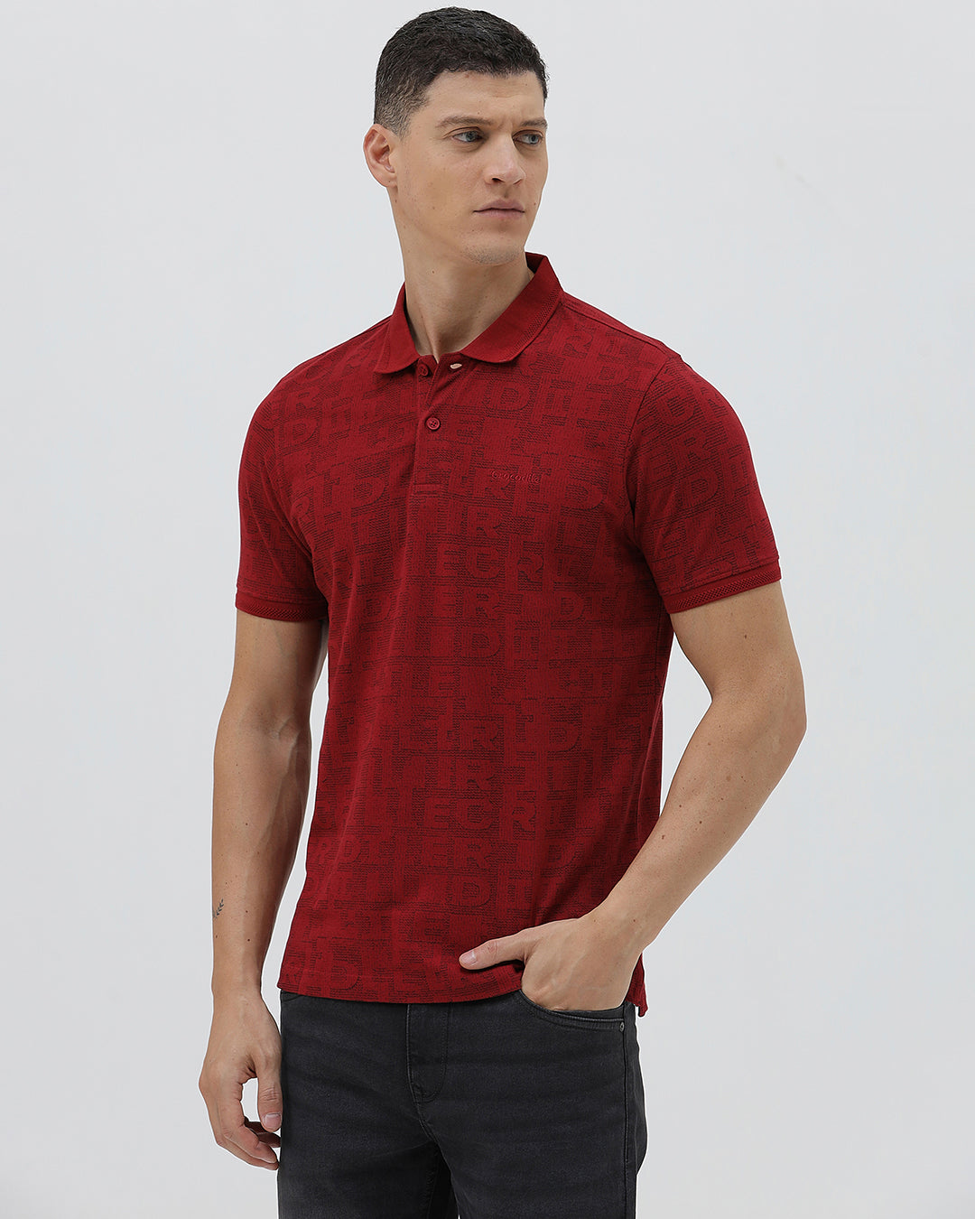 A man wearing a red short-sleeve polo shirt with a subtle tonal monogram pattern, paired with dark gray jeans, standing against a light gray studio background.