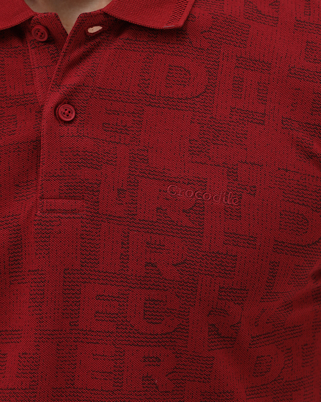 A man wearing a red short-sleeve polo shirt with a subtle tonal monogram pattern, paired with dark gray jeans, standing against a light gray studio background.
