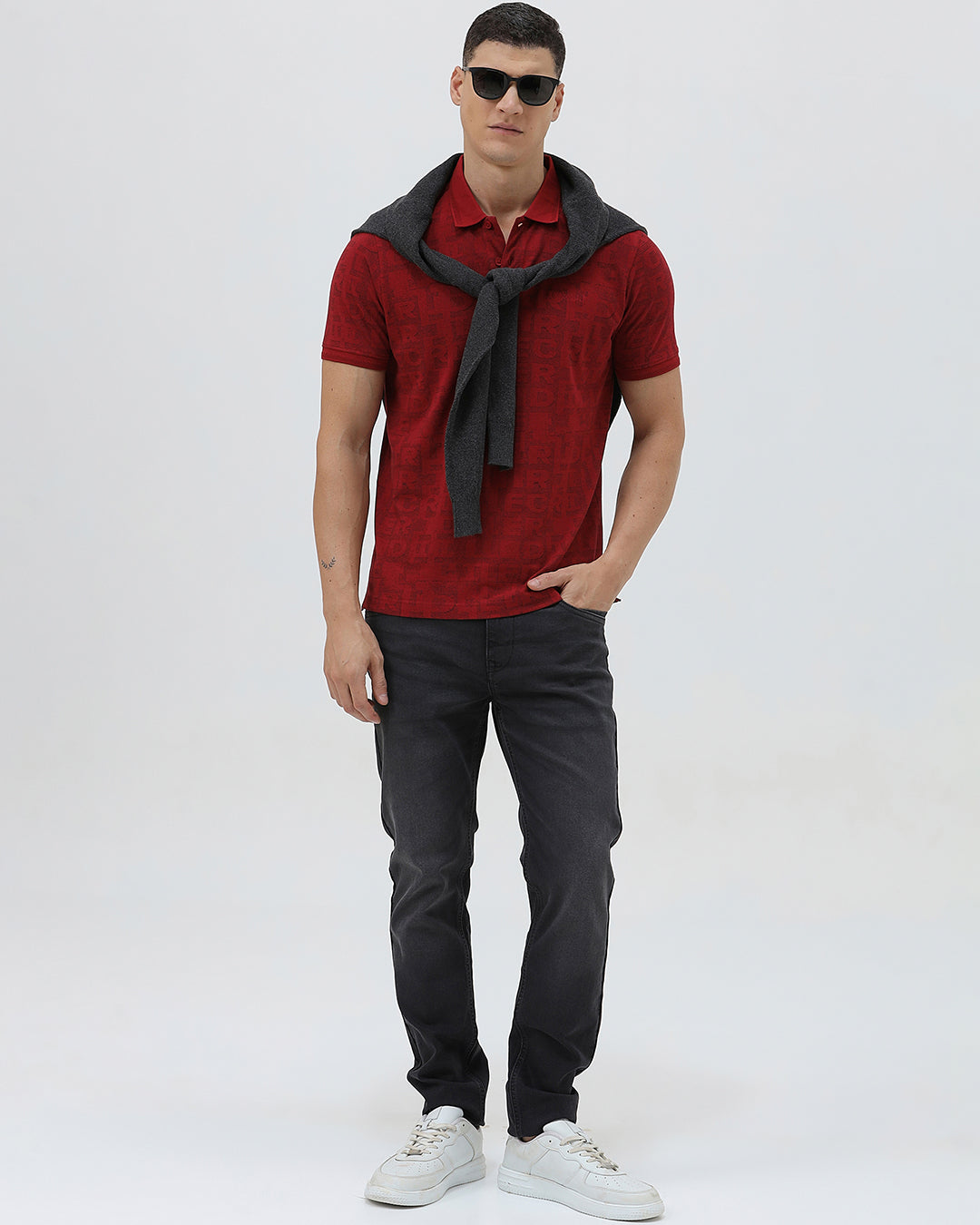 A man wearing a red short-sleeve polo shirt with a subtle tonal monogram pattern, paired with dark gray jeans, standing against a light gray studio background.