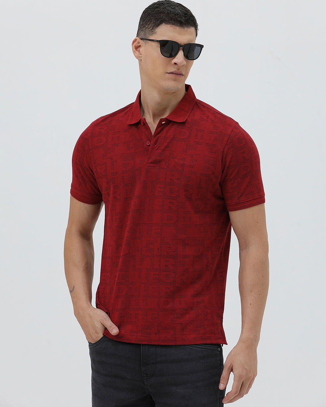 A man wearing a red short-sleeve polo shirt with a subtle tonal monogram pattern, paired with dark gray jeans, standing against a light gray studio background.