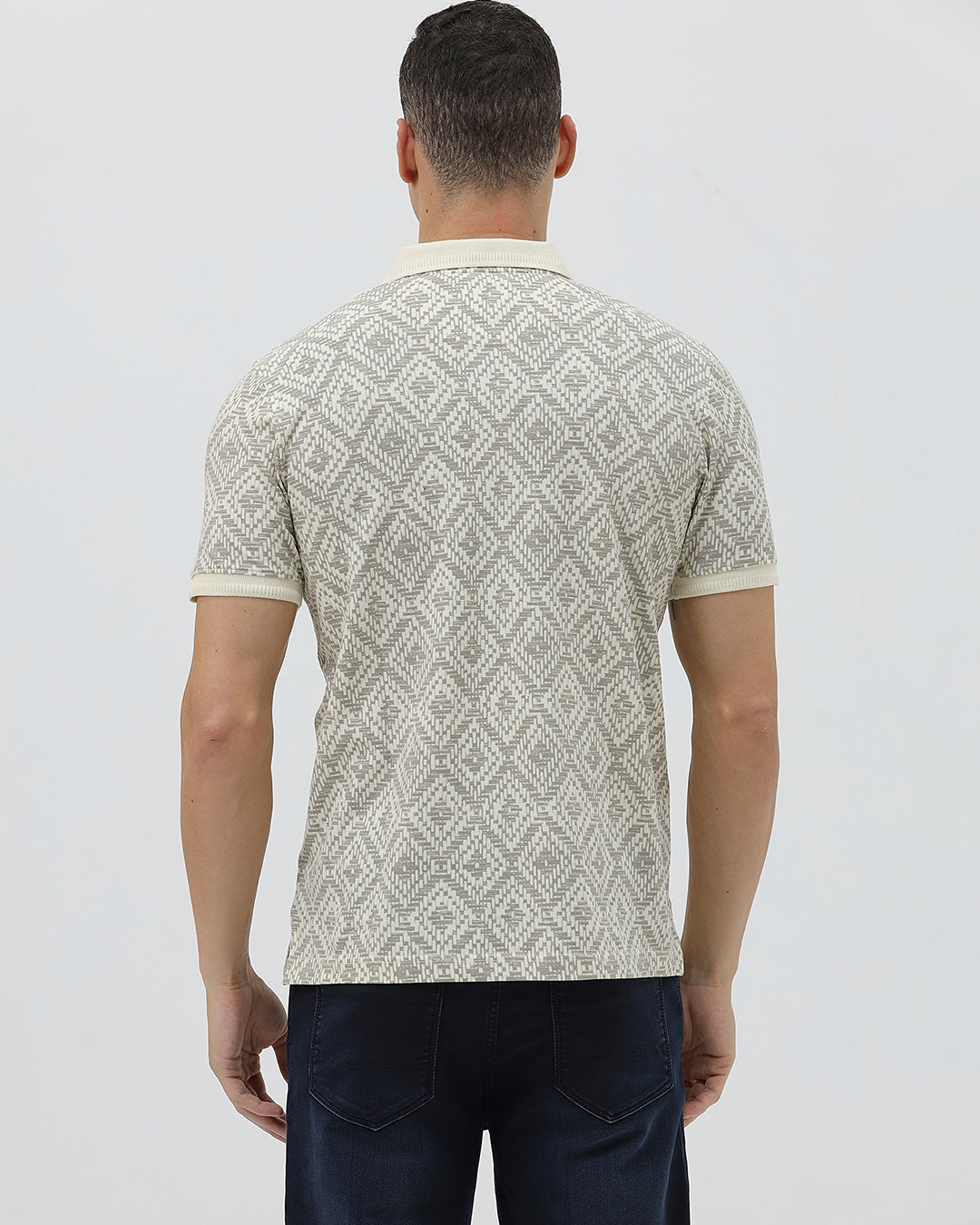 A man wearing a short-sleeve polo shirt with a beige and gray geometric pattern, paired with dark blue jeans, standing against a light gray background.